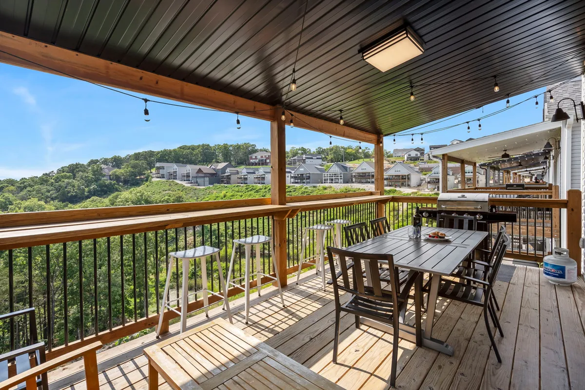 A scenic outdoor deck featuring a wooden dining table, barbecue grill, and panoramic views of hillside houses. The relaxed, social setting invites guests to gather, dine, and take in the beautiful surroundings, linking the home to Branson’s appealing local environment.