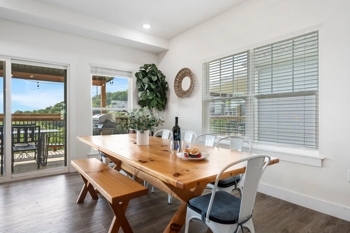 A warmly lit dining area featuring a wooden table set for a meal, complemented by a green plant and decorative elements. A bottle of wine and plates of food set the stage for convivial gatherings, with glimpses of scenic views through nearby windows.