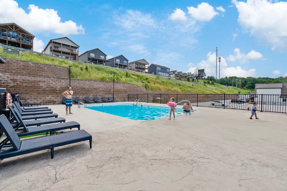 A sunlit outdoor swimming pool with families and children enjoying the water, surrounded by lounge chairs. The joyful atmosphere is enhanced by the presence of people relaxing and playing, suggesting a vibrant, community-focused amenity area.