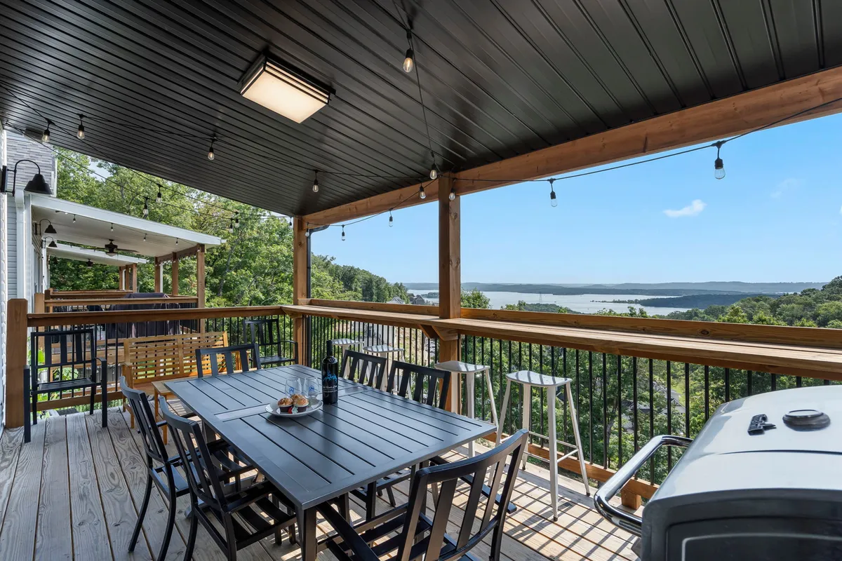 An exterior dining table set up on a deck, with string lights above, a grill nearby, and a view of the water in the background. The ambiance is relaxed and festive, perfect for family meals or casual gatherings as the sun sets.