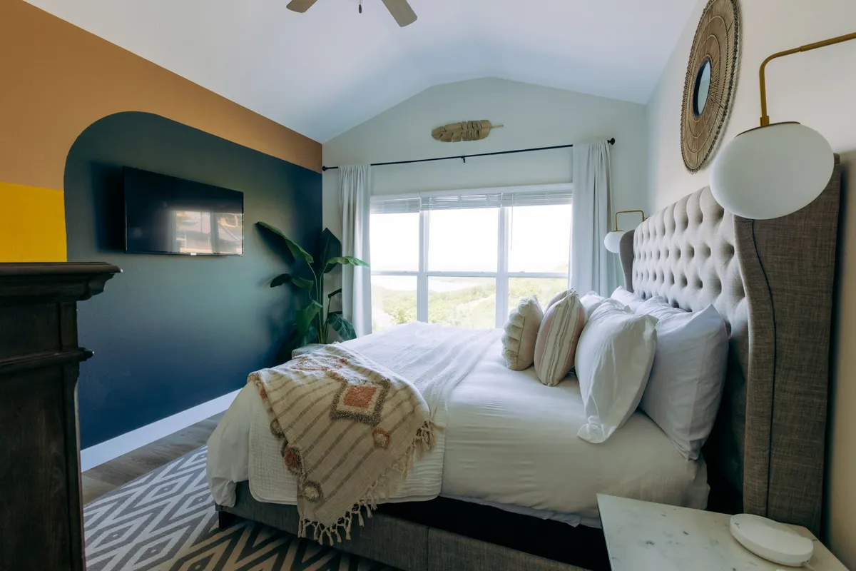 A bright and tranquil bedroom with a large bed, window view, stylish wall decor, a television, and a green plant. The space feels airy and peaceful, perfect for unwinding after a busy day. The arrangement and features suggest a well-appointed, comfortable sanctuary.