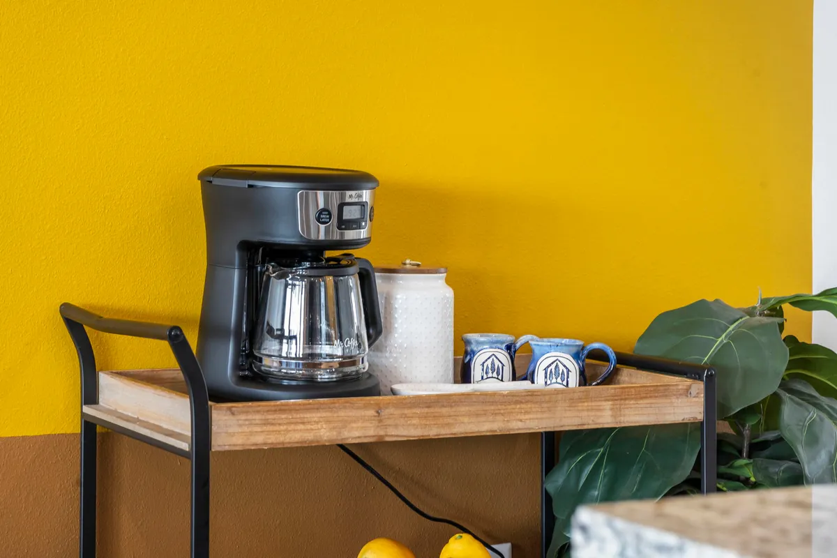 A close-up of a cozy coffee station featuring a coffee maker, an array of mugs, a cheerful yellow wall, and a wooden cart adorned with a leafy plant. The setup is both practical and visually inviting, ready for morning rituals.