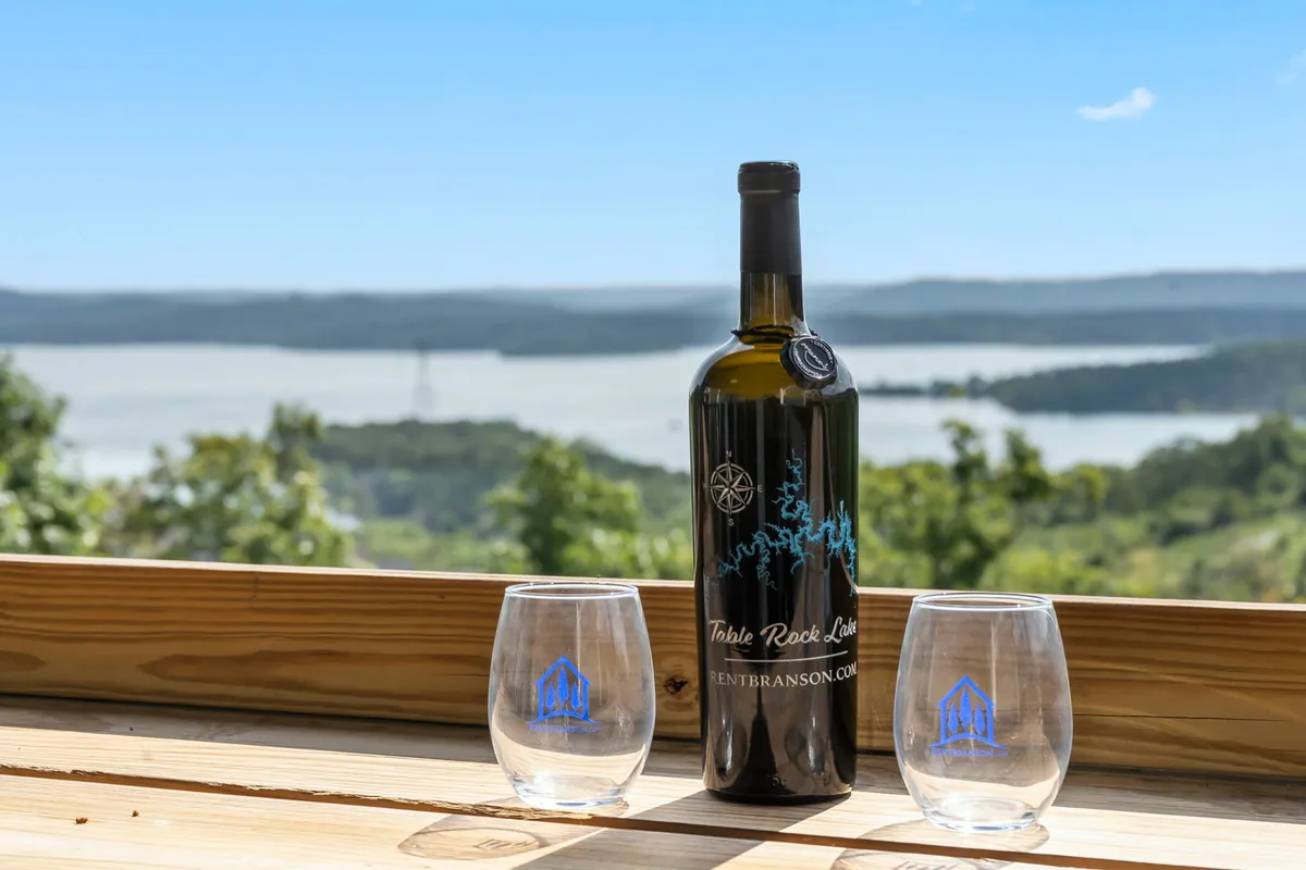 This image captures a bottle of wine and two glasses perched on an outdoor table, with a scenic view stretching out behind. The setting sun lights up the horizon, creating a romantic and relaxed atmosphere perfect for couples or friends to enjoy together.