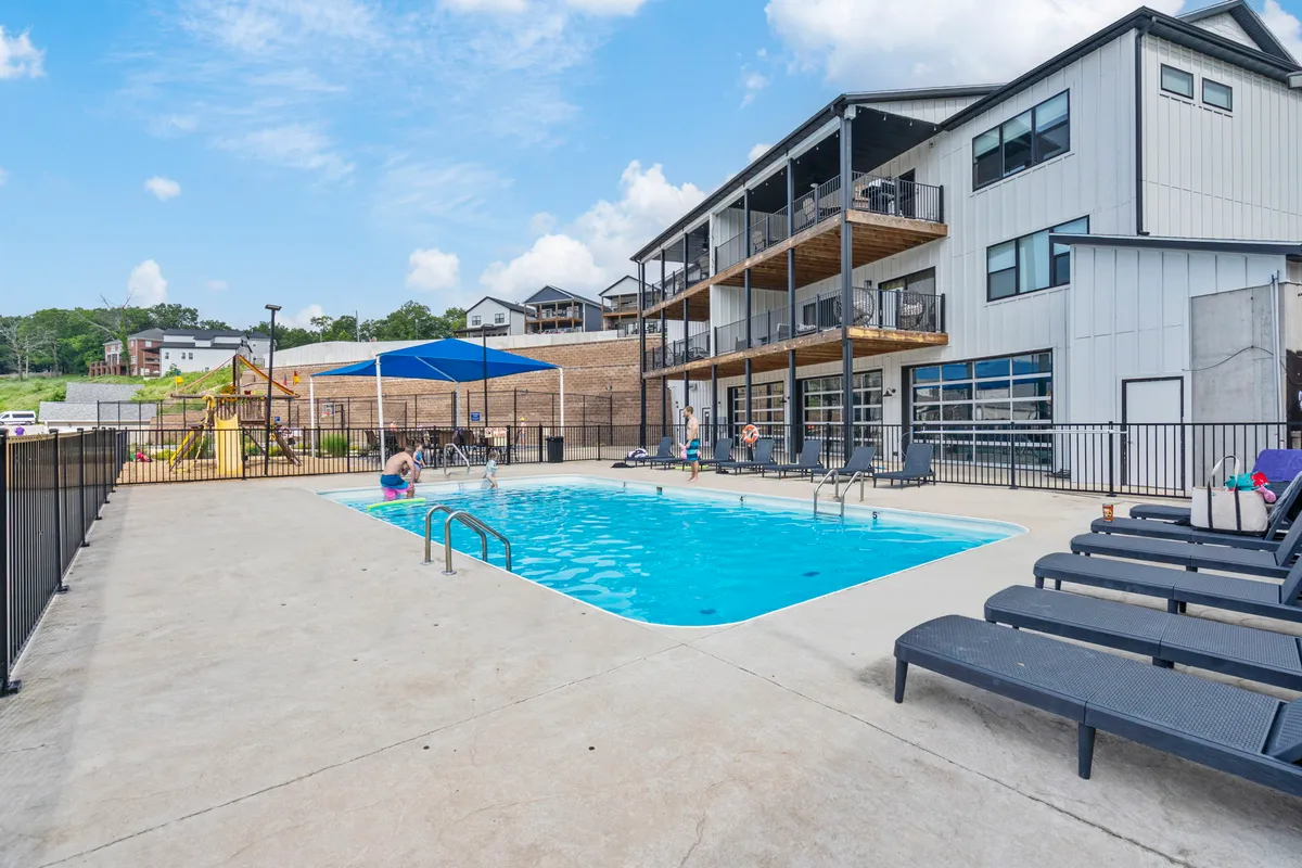 A lively scene featuring an exterior swimming pool surrounded by lounge chairs, a nearby playground area, and the facade of the main building. Children are playing, and people are socializing, creating an energetic and communal vibe.