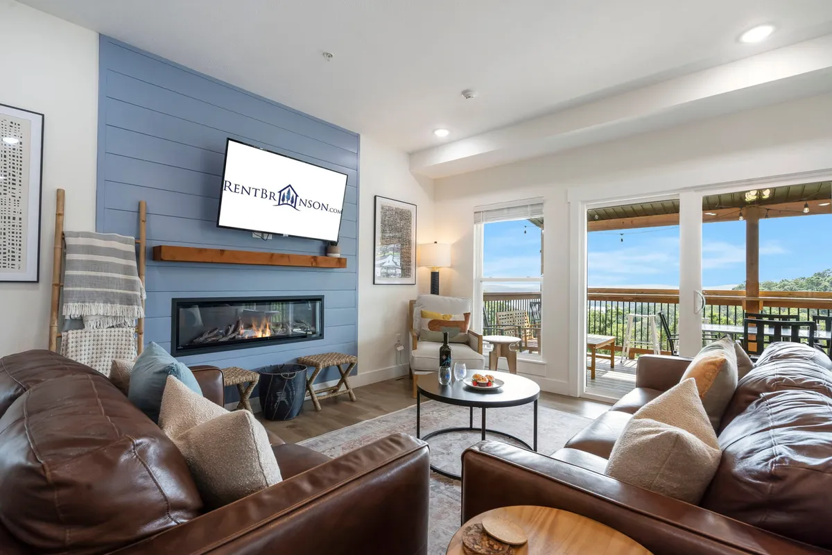 A cozy living room with leather sofas arranged around a modern fireplace, a large television, and expansive windows framing serene lake views. The space glows with natural light, creating an inviting atmosphere ideal for unwinding with friends or family.