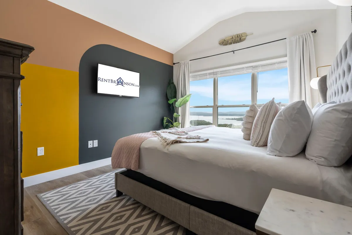 A cozy bedroom featuring a comfortable bed, a television, a window that offers a view, and a calming art piece. The decor is soothing, with soft bedding and neutral tones that foster relaxation. The window hints at natural light and potentially a serene outside view, adding to the restful atmosphere.